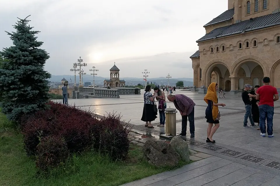 Прихожане, некоторые в платках, собираются в сумерках на каменном дворе у собора Самеба