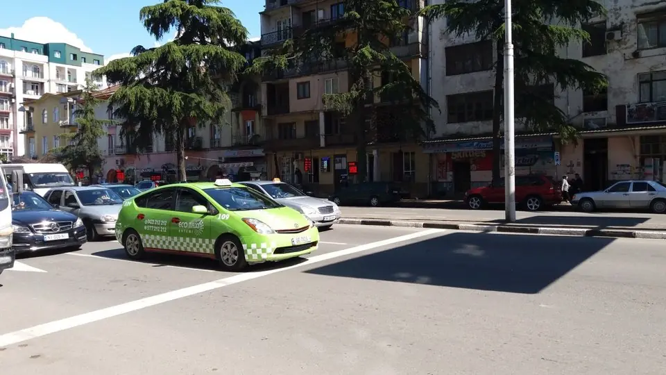 A lime-green Eco Taxi Toyota Prius with a roof sign, waiting on Chavchavadze Street in Batumi beside tall cedar trees