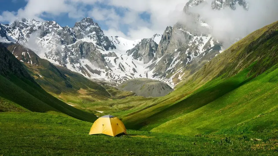 Yellow tent in a green valley below jagged snowy peaks and clouds