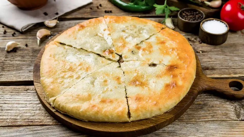 A whole kubdari cut into wedges on a wooden board, with garlic, peppercorns and a bowl of salt beside it
