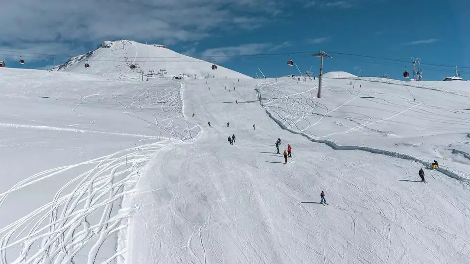 Skiers on a wide groomed piste at Gudauri under a chairlift, with snowy Caucasus ridges behind