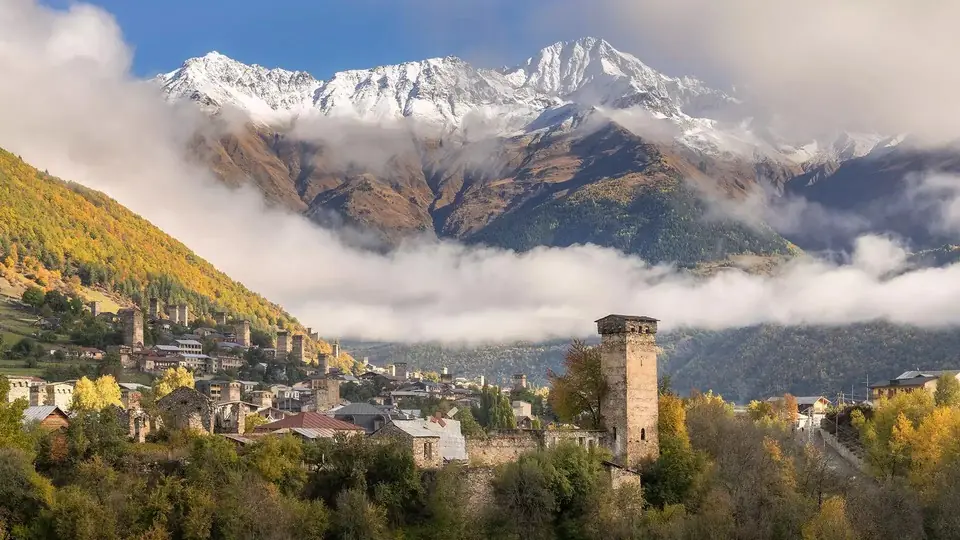 Mestia stone towers and houses below a band of cloud and snowy Caucasus peaks in autumn