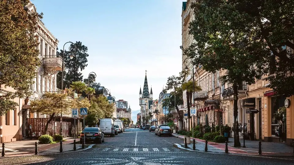 A cobbled street in Old Batumi lined with pastel 19th-century houses, palms and plane trees, leading to the spired clock tower on Europe Square
