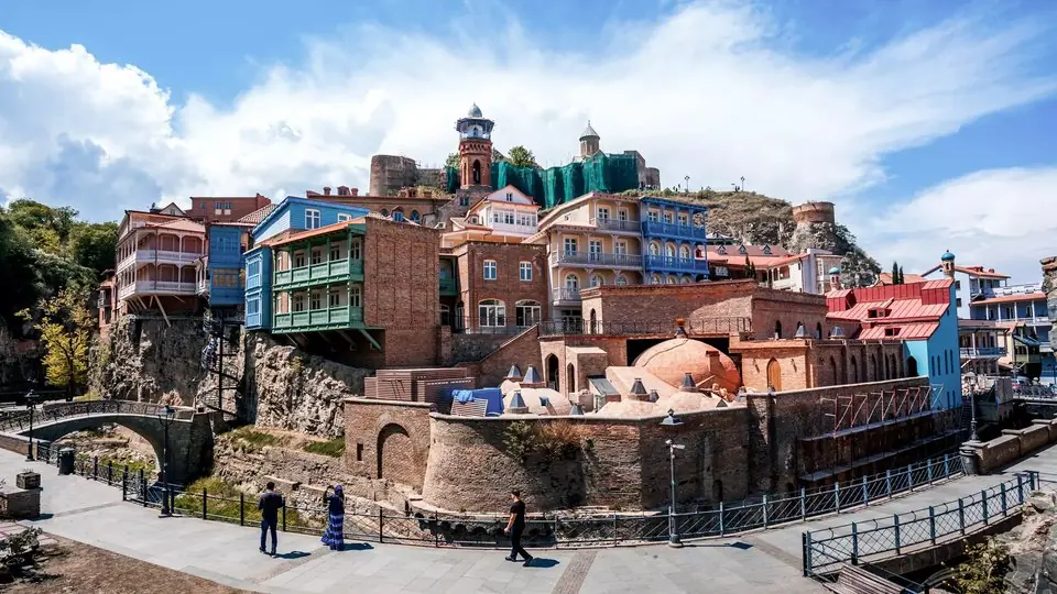 Balconied old houses in green, blue and red stacked above the brick domes of the Abanotubani sulphur baths, with a mosque minaret and Narikala fortress behind