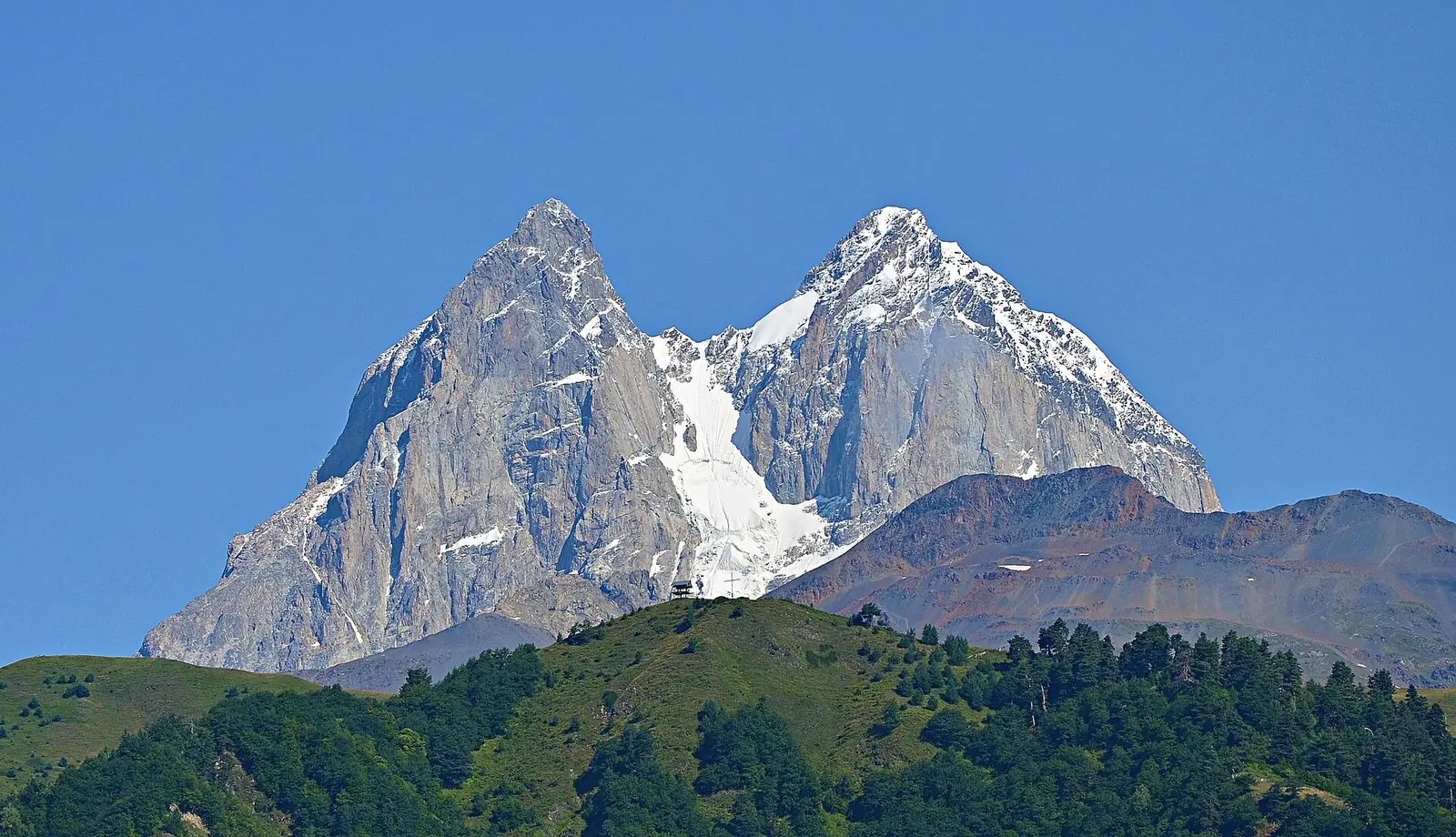 The two granite summits of Mount Ushba with a snow-filled couloir between them, rising above a forested ridge in Svaneti