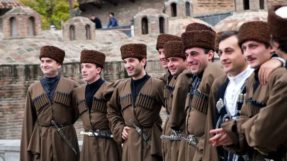 Ряд улыбающихся фольклорных артистов в коричневых чохах на фоне куполов серных бань Тбилиси