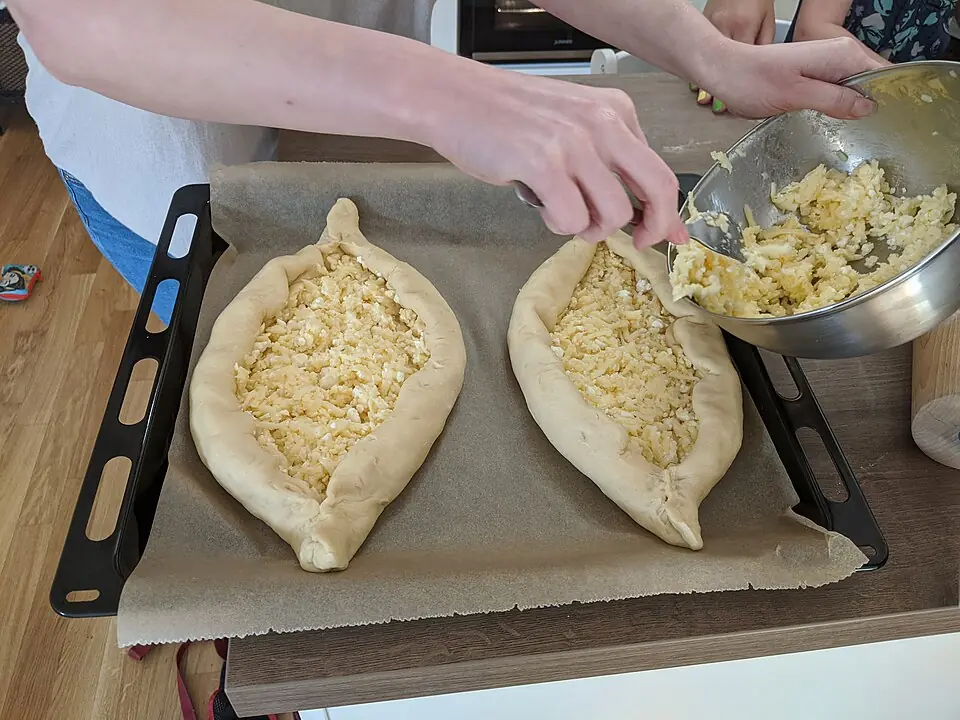 Hands spooning grated cheese filling into two boat-shaped khachapuri dough cases on a lined baking tray