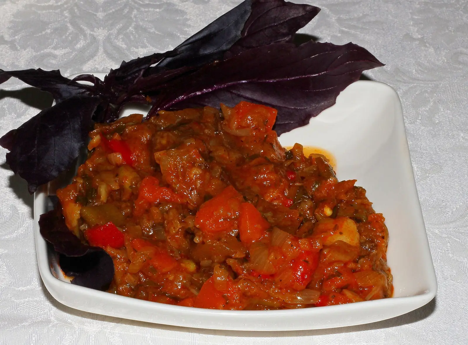 Ajapsandali in a white square dish, a thick stew of aubergine, red pepper, tomato and onion, with sprigs of purple basil tucked behind it