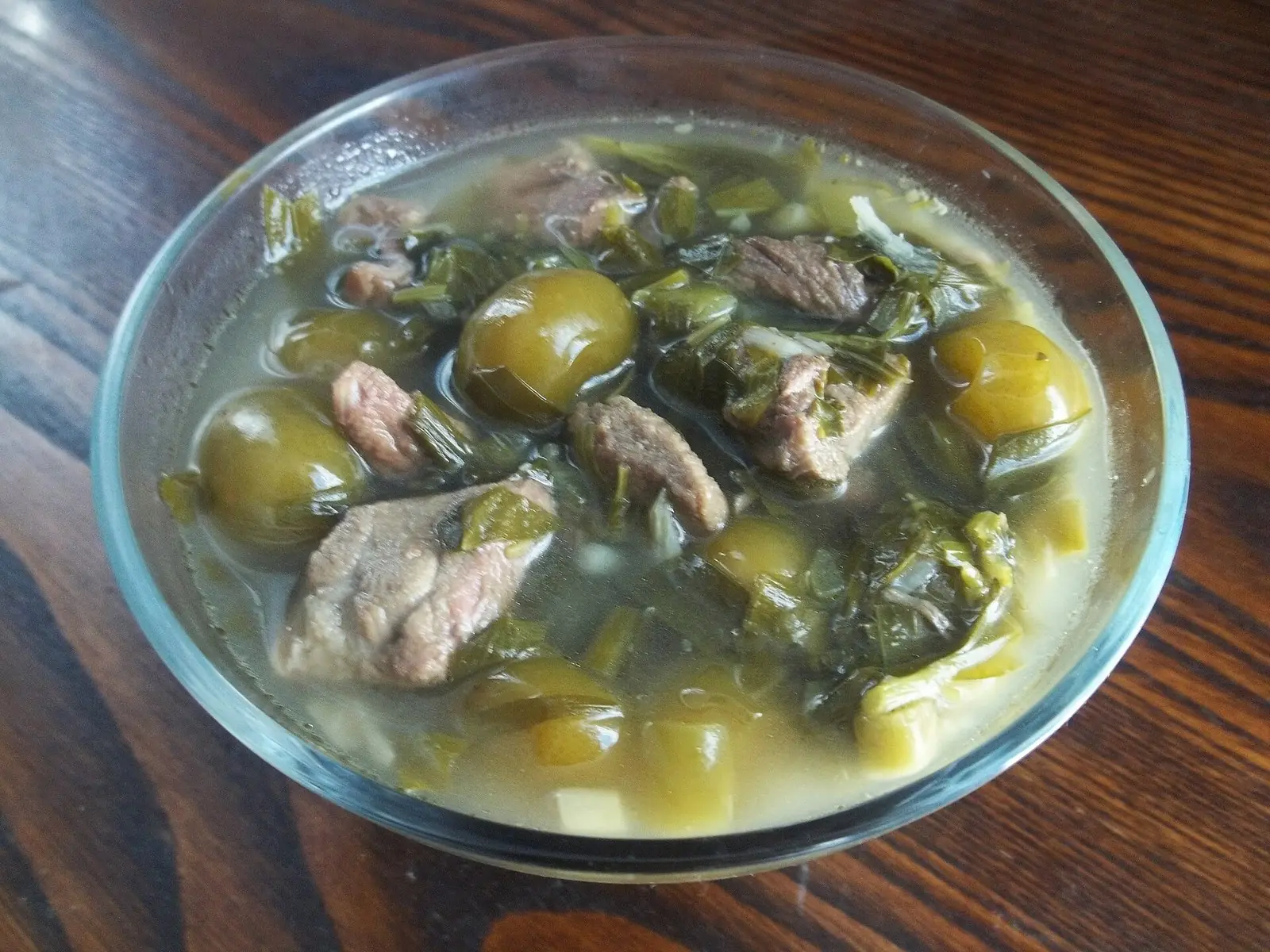 Chakapuli in a glass bowl on a wooden table: pieces of lamb and whole green plums in a pale broth full of wilted tarragon and spring onion
