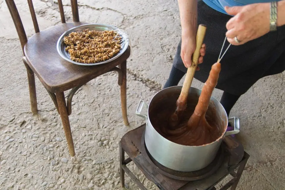A string of walnuts lifted out of a pot of thick grape-juice batter (tatara) on an outdoor stove, with more strings of walnuts waiting on a chair