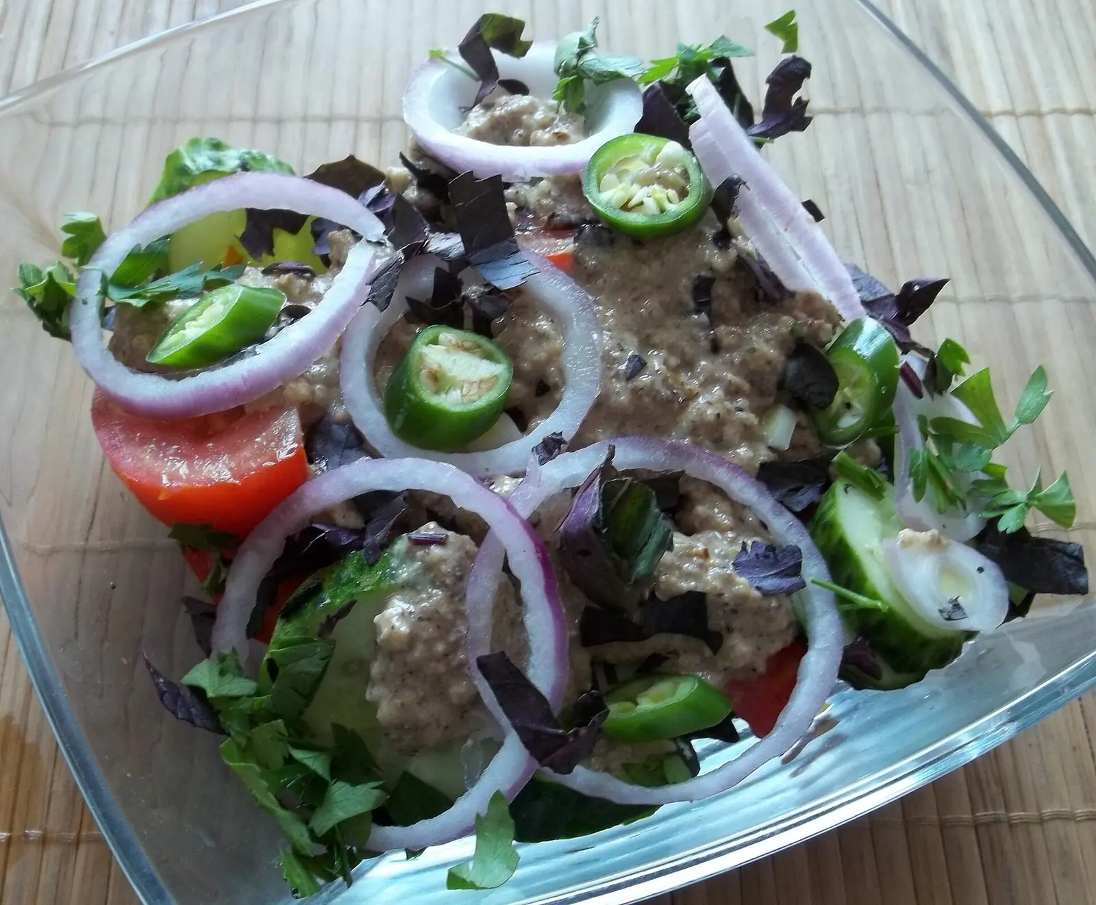 Georgian salad with walnuts in a glass bowl: tomato and cucumber under a creamy walnut dressing, topped with red onion rings, sliced green chilli, purple basil and coriander