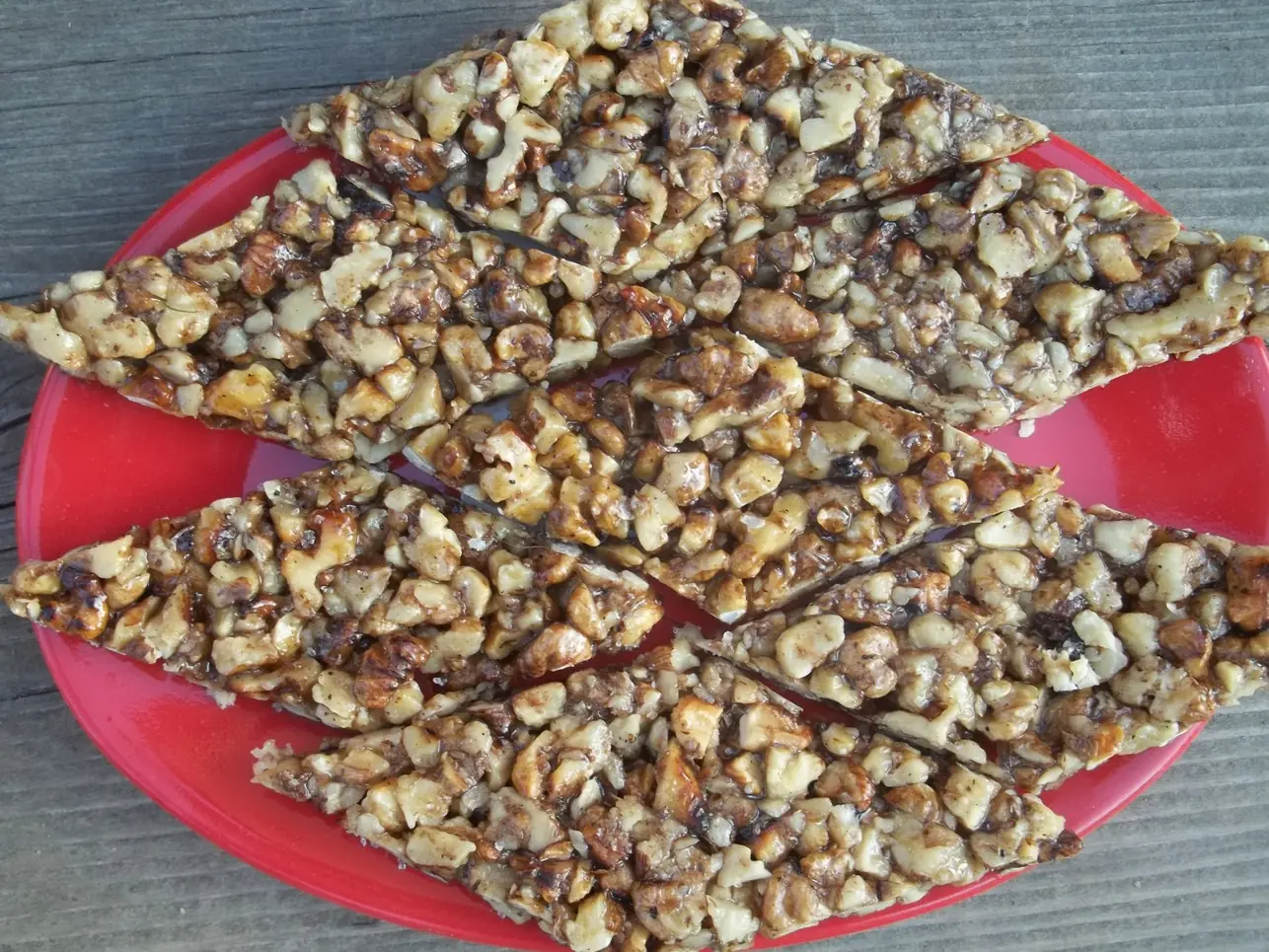 Long diamond-shaped pieces of homemade walnut gozinaki, glossy with set honey, laid on a red plate on a weathered wooden table