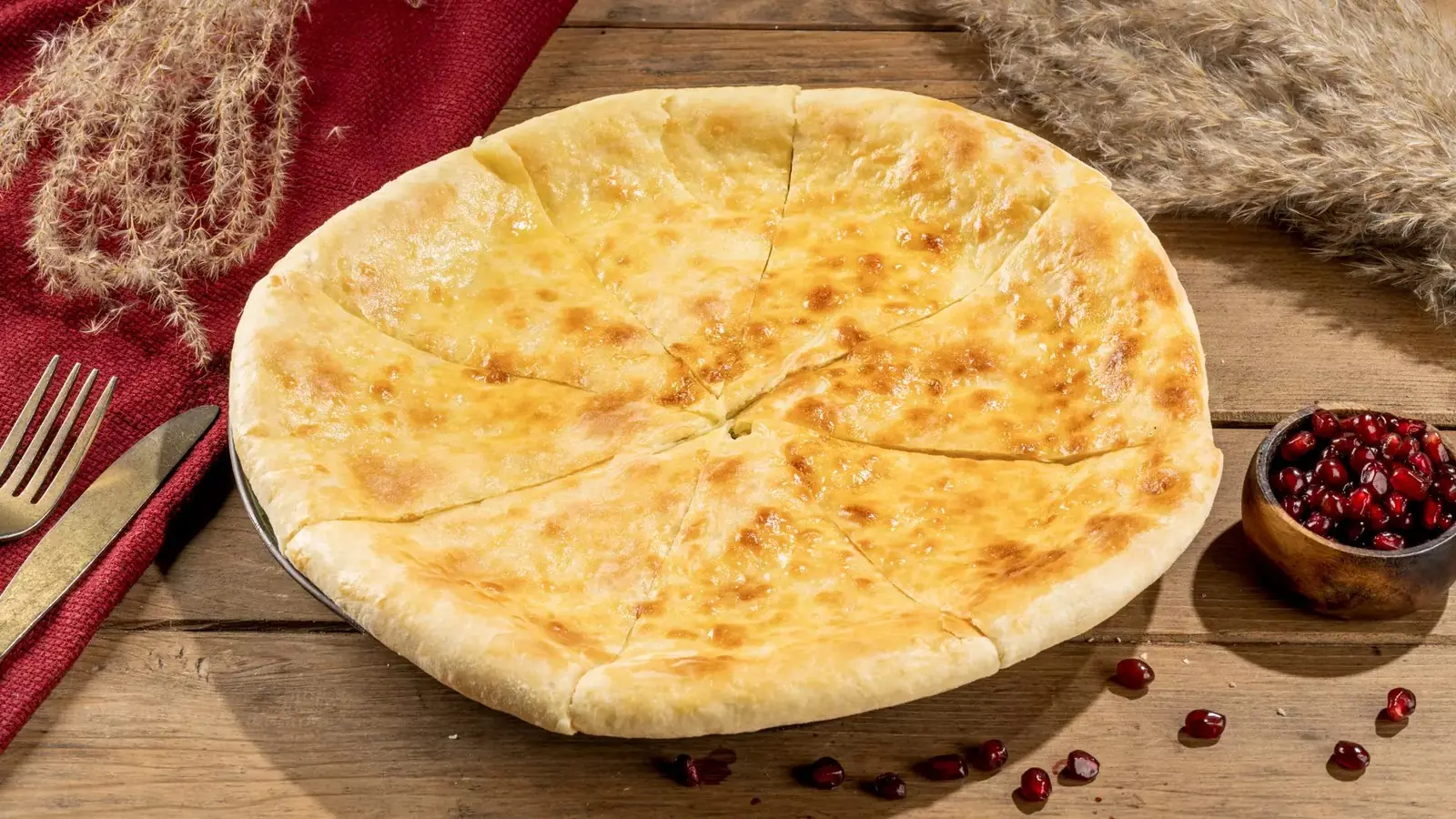 A golden round Imeruli khachapuri cut into eight wedges on a wooden table, with a red cloth, dried grasses and a small bowl of pomegranate seeds
