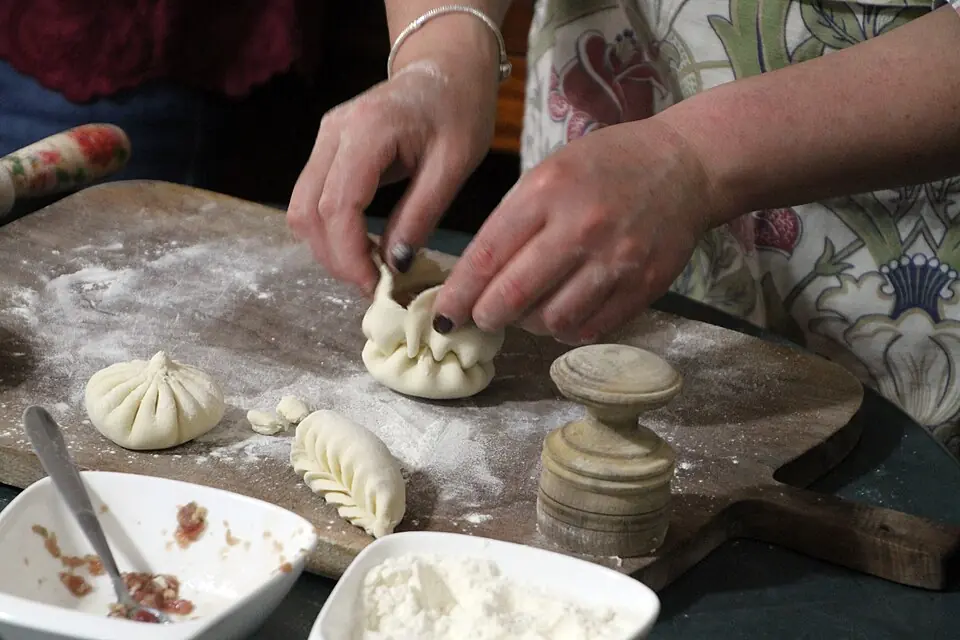 Hands pleating a khinkali dumpling on a floured wooden board, with a finished dumpling, a bowl of meat filling and a bowl of flour beside it
