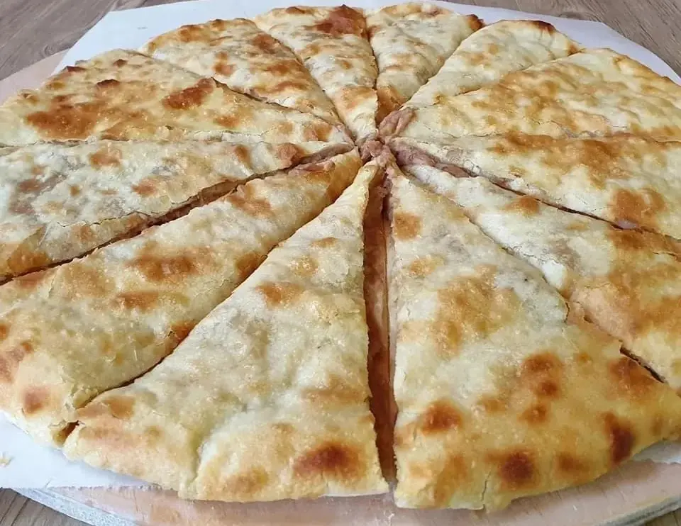 A round homemade lobiani cut into wedges on baking paper, the mashed bean filling showing between pale golden, brown-spotted pieces of crust