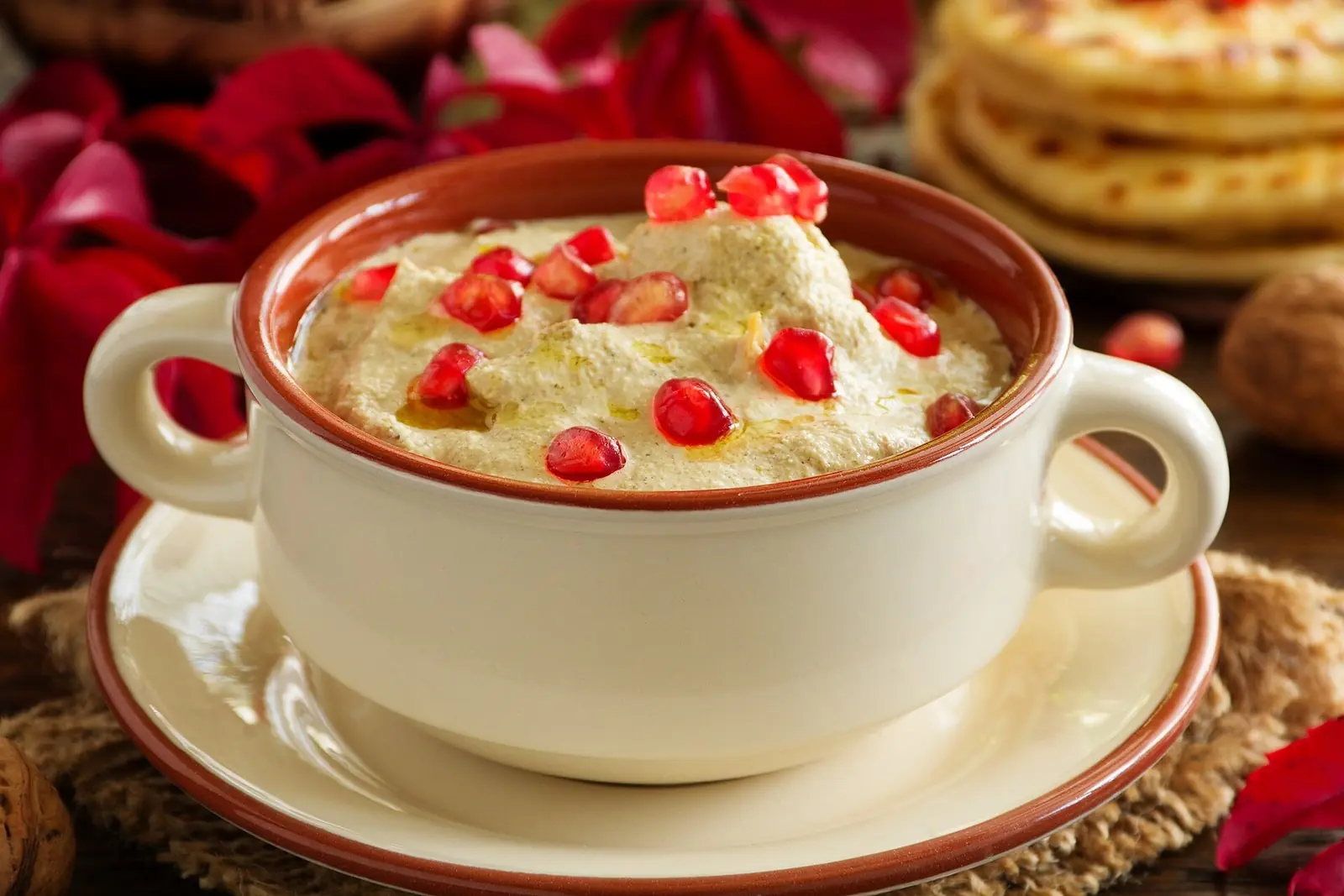 A cream two-handled bowl of satsivi on a matching saucer, the thick walnut sauce glossed with walnut oil and scattered with pomegranate seeds, with khachapuri, whole walnuts and red leaves behind
