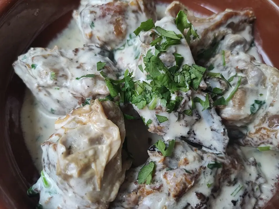 Close-up of shkmeruli: fried chicken pieces in a creamy garlic sauce in a clay dish, sprinkled with chopped herbs