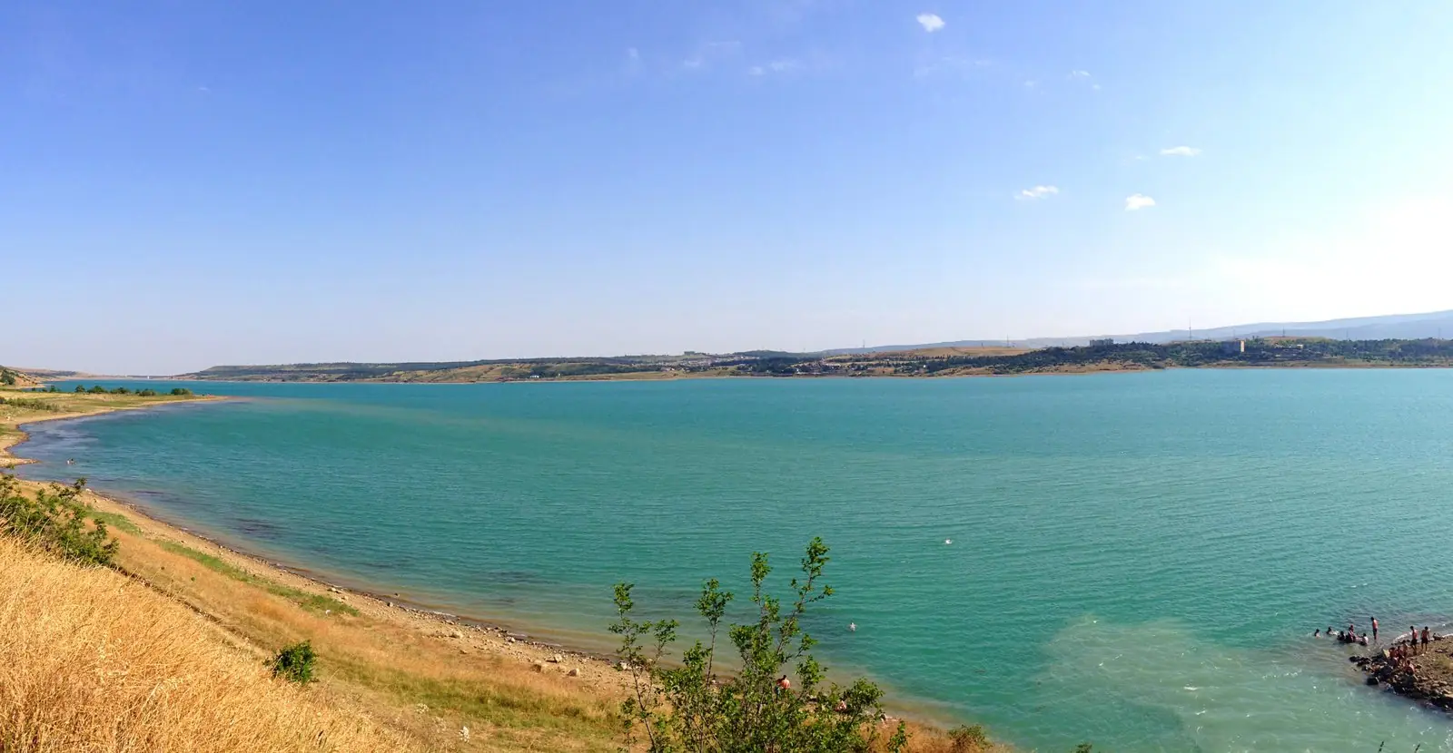 Turquoise water of the Tbilisi Sea below a dry grassy bank, with swimmers on the rocks at the right and the far shore on the horizon