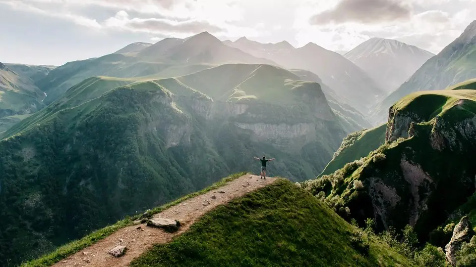 Hiker with arms raised at the end of a ridge path above deep green Kazbegi gorges