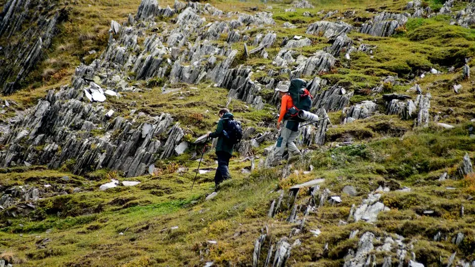 Two trekkers with big backpacks crossing a slope of jagged slate rocks in Khevsureti