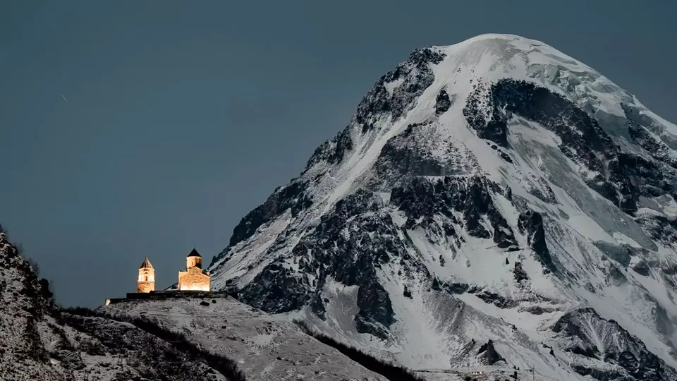 Подсвеченная Троицкая церковь в Гергети в сумерках под снежным куполом Казбека