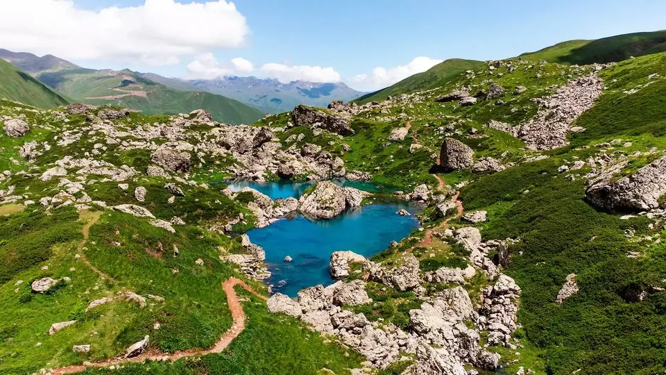 Turquoise Abudelauri lake among boulders and green slopes, with a dirt hiking path winding past it