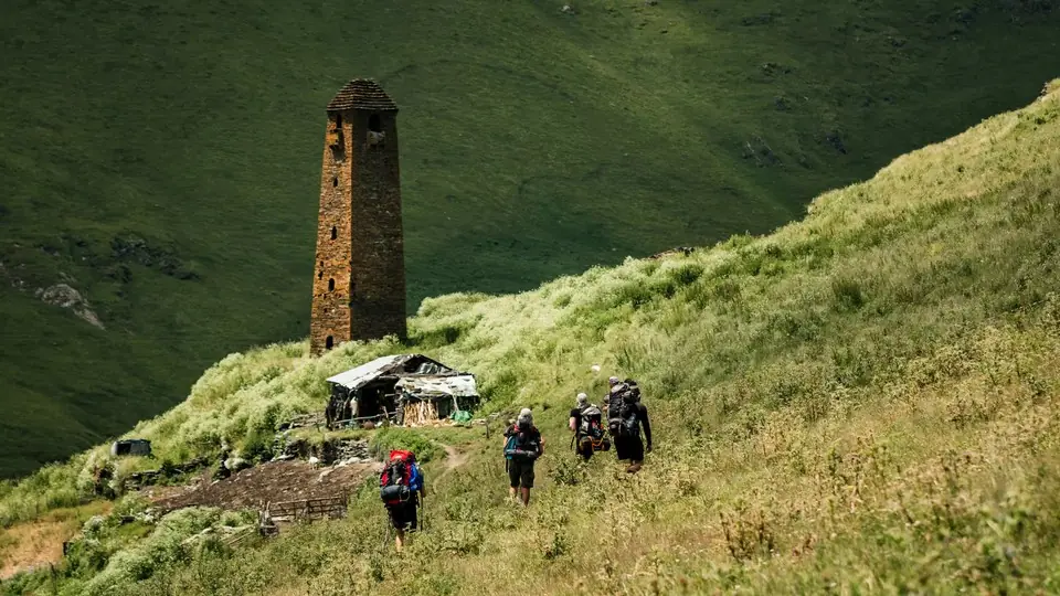 Backpackers walking up a grassy slope past an old stone tower and a shepherd hut