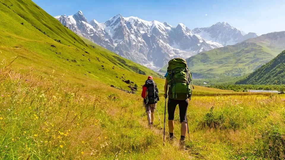 Two backpackers walking through a flowery meadow towards the snowy Shkhara wall